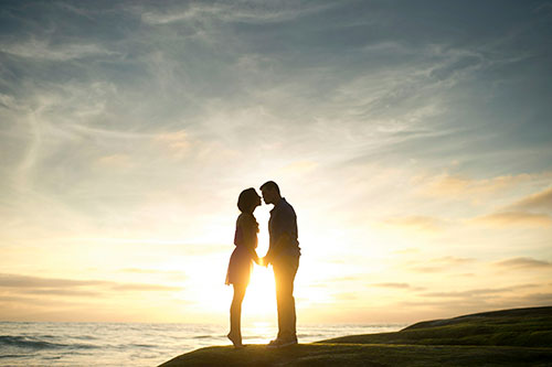 Silhouette of a couple holding hands against a sunset backdrop by the ocean.